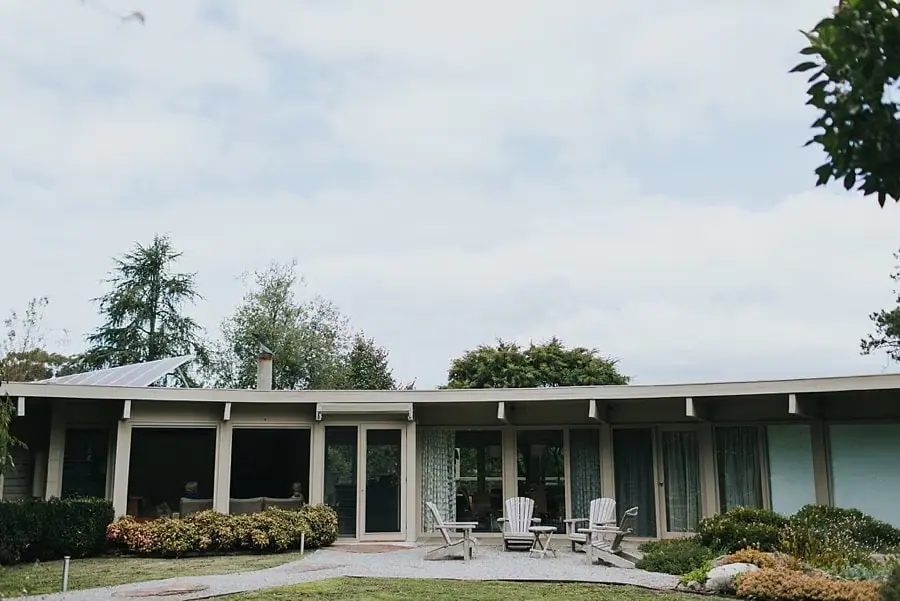 Low modern house with glass doors and outdoor chairs beneath a cloudy sky.