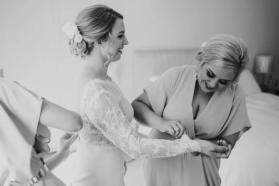 Black-and-white photograph of the smiling bride having her sleeve adjusted by a companion.