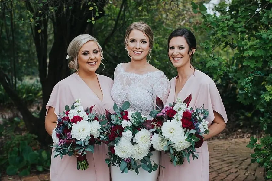 Bride and two women in pink dresses posing with red and white bouquets in a garden.