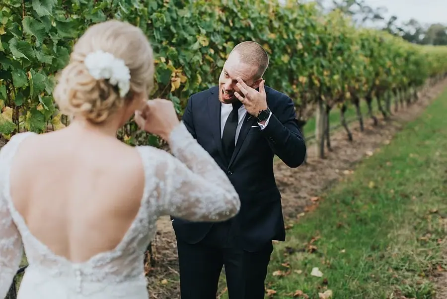 Groom wiping his eye while the bride stands opposite him among grapevines.