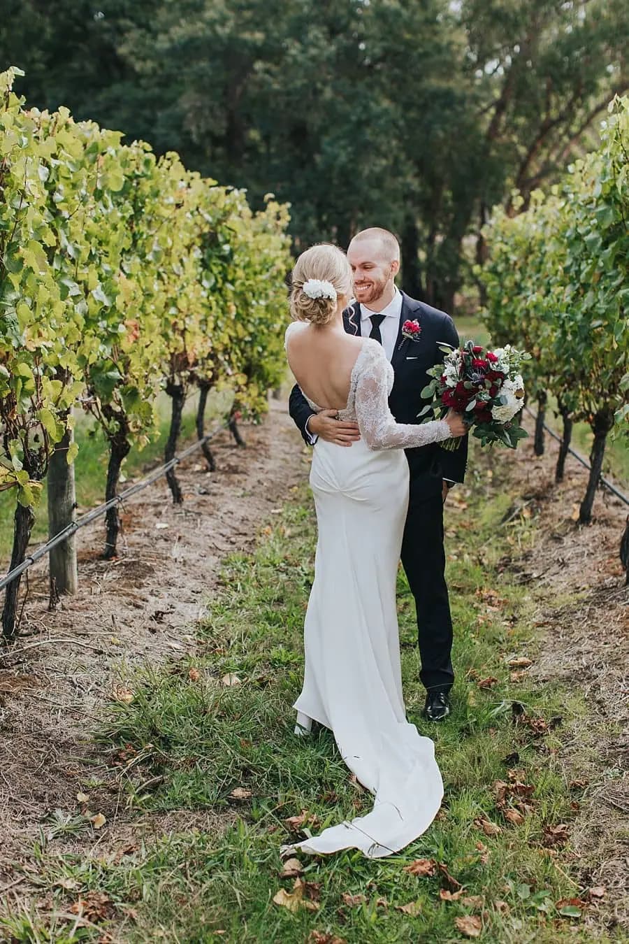 Bride and groom embracing on a path between vineyard rows.