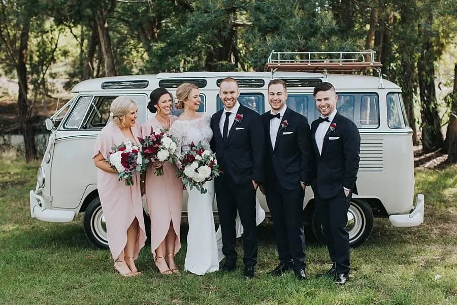 Wedding party posing beside a white vintage Volkswagen van beneath leafy trees.