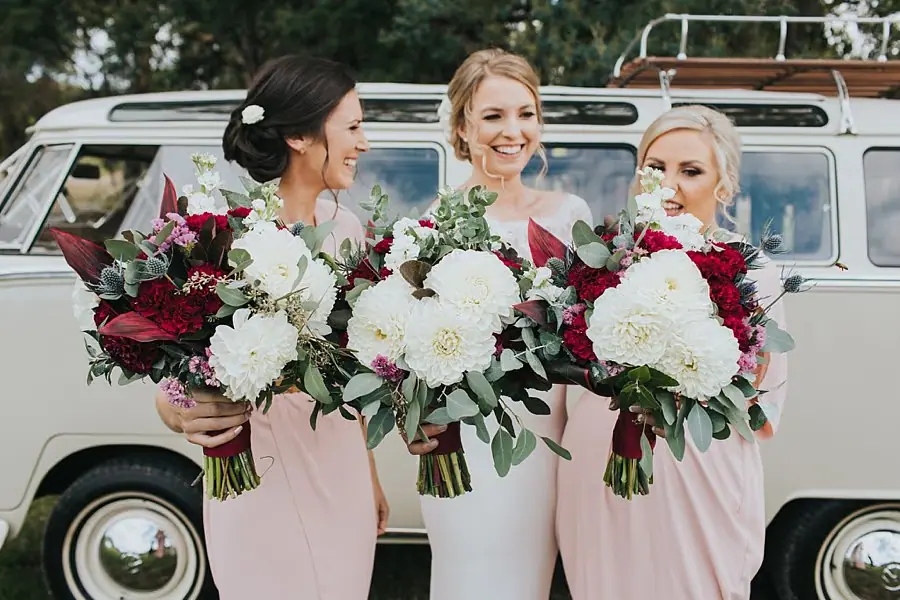 Bride and two women smiling with red and white bouquets in front of the van.