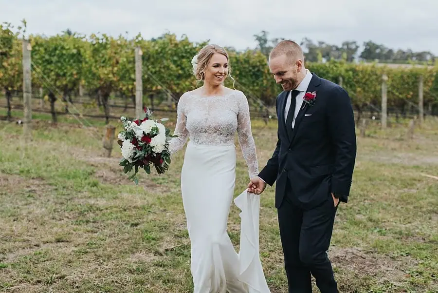 Bride and groom smiling and holding hands while walking beside grapevines.