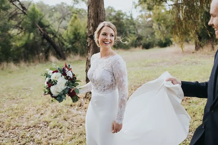 Bride smiling and holding her bouquet while the groom lifts her dress train.