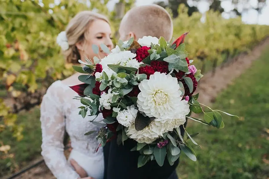 Red and white bouquet in the foreground as the couple lean close behind it.