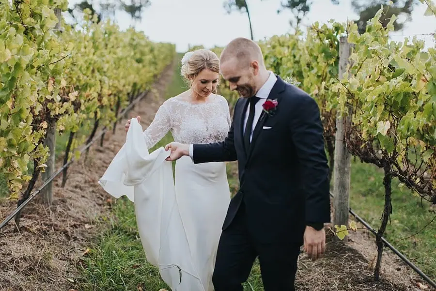 Bride and groom looking down as they walk together between grapevines.