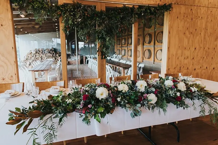 Long floral arrangement across a white reception table beneath hanging greenery.