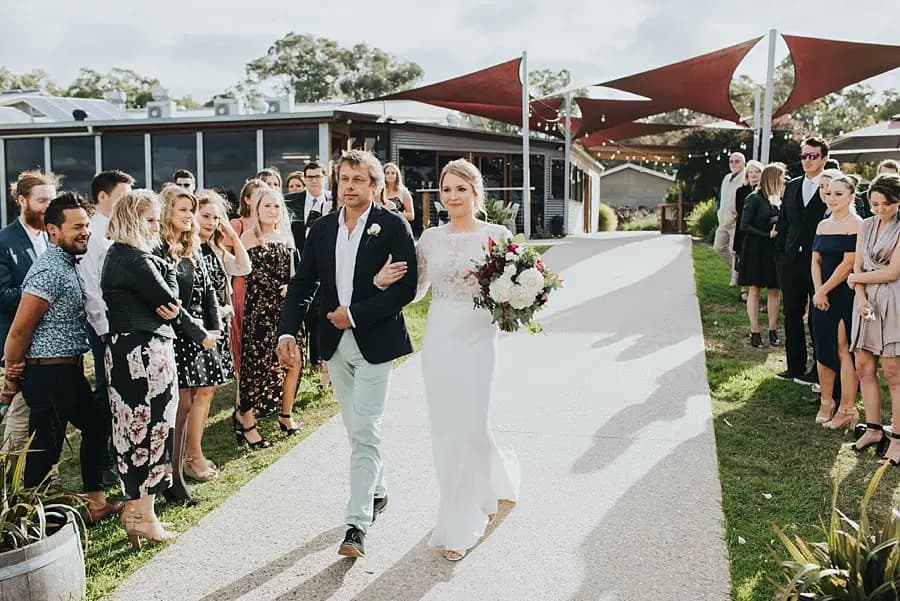 Bride walking down the aisle with a man in a dark jacket and pale trousers.