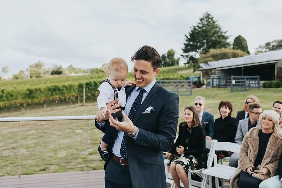 Man holding a small child wearing a bow tie beside seated ceremony guests.