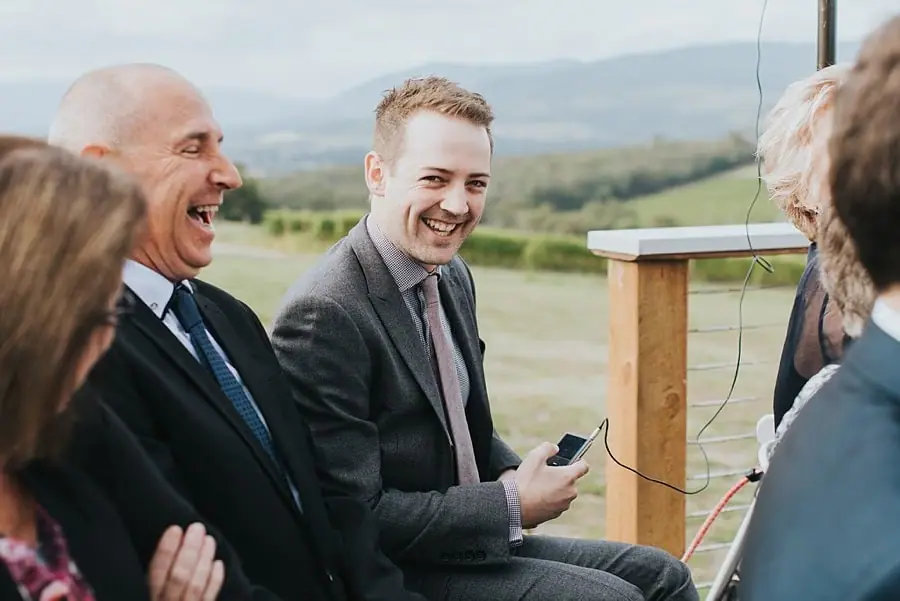 Two men laughing among seated guests outside the ceremony.