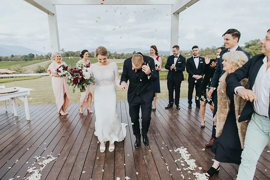 Couple walking down the wooden aisle as guests throw white petals.