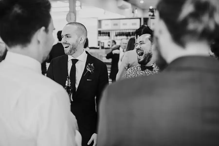 Black-and-white photograph of the groom laughing with guests indoors.