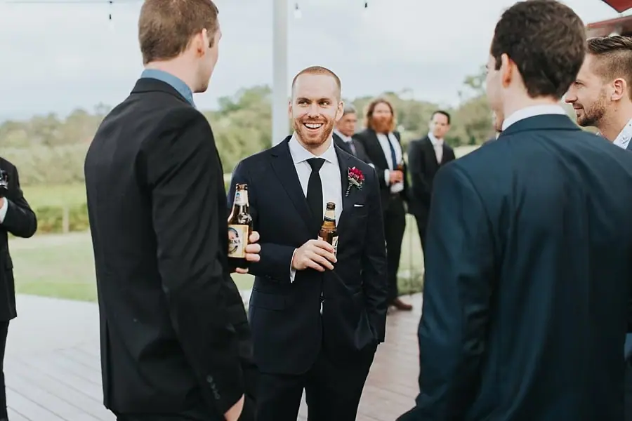 Groom laughing with men holding bottles beside the ceremony deck.