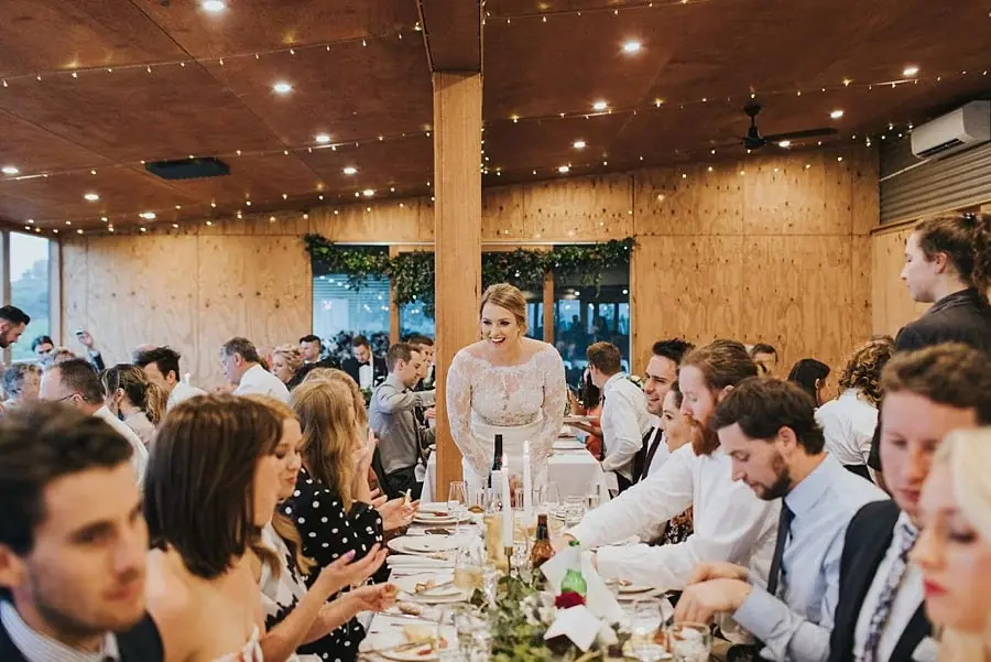 Bride standing and smiling among guests at a long reception table.