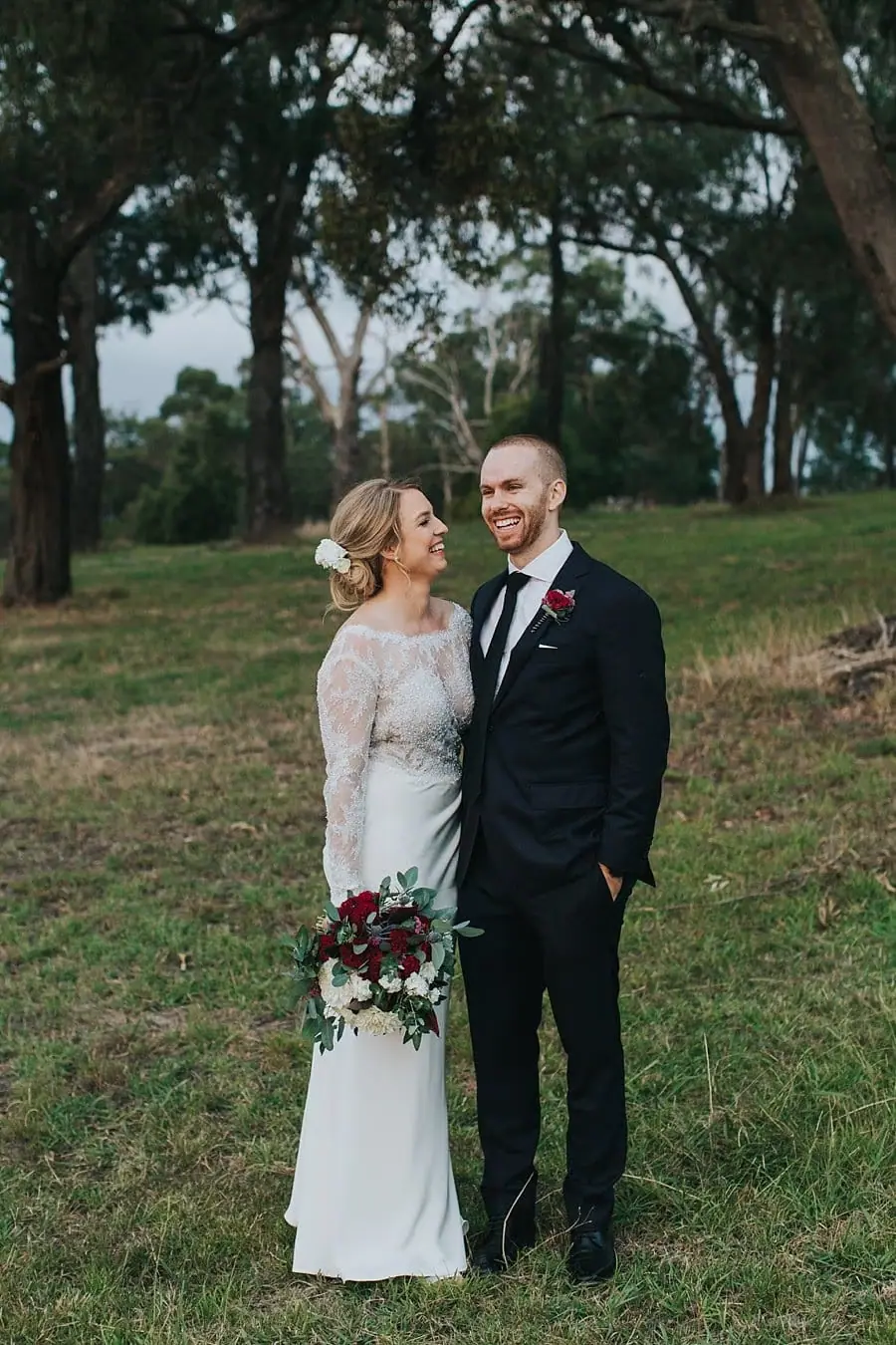 Bride and groom standing together beneath leafy trees with her bouquet.