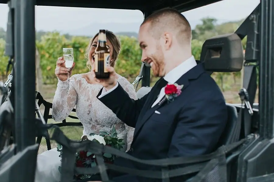 Bride and groom raising drinks while sitting together in a small open vehicle.