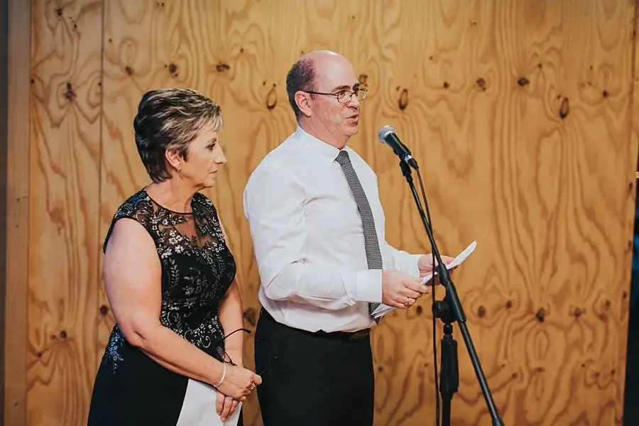 Man reading a microphone speech beside a woman against a timber wall.