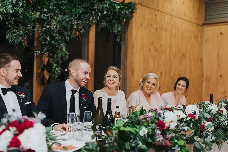 Bride and groom laughing with their wedding party behind a floral table arrangement.