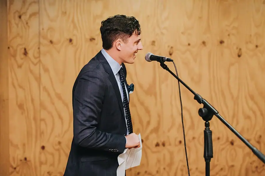 Man giving a microphone speech against a pale timber wall.