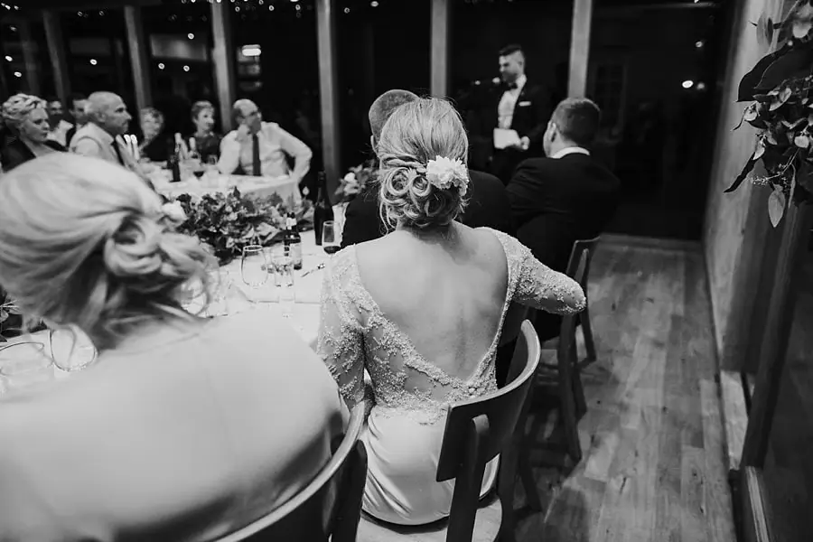 Black-and-white photograph of the bride listening to a speech from her reception chair.