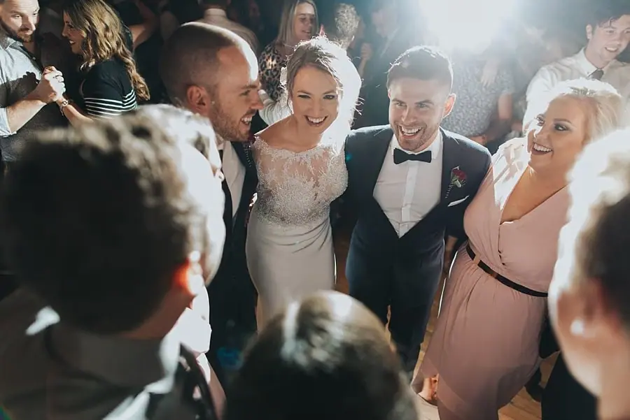 Bride and groom laughing with their wedding party on the crowded dance floor.