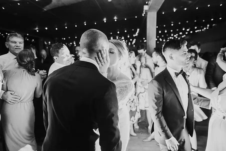 Black-and-white photograph of the bride and groom kissing among dancing guests.