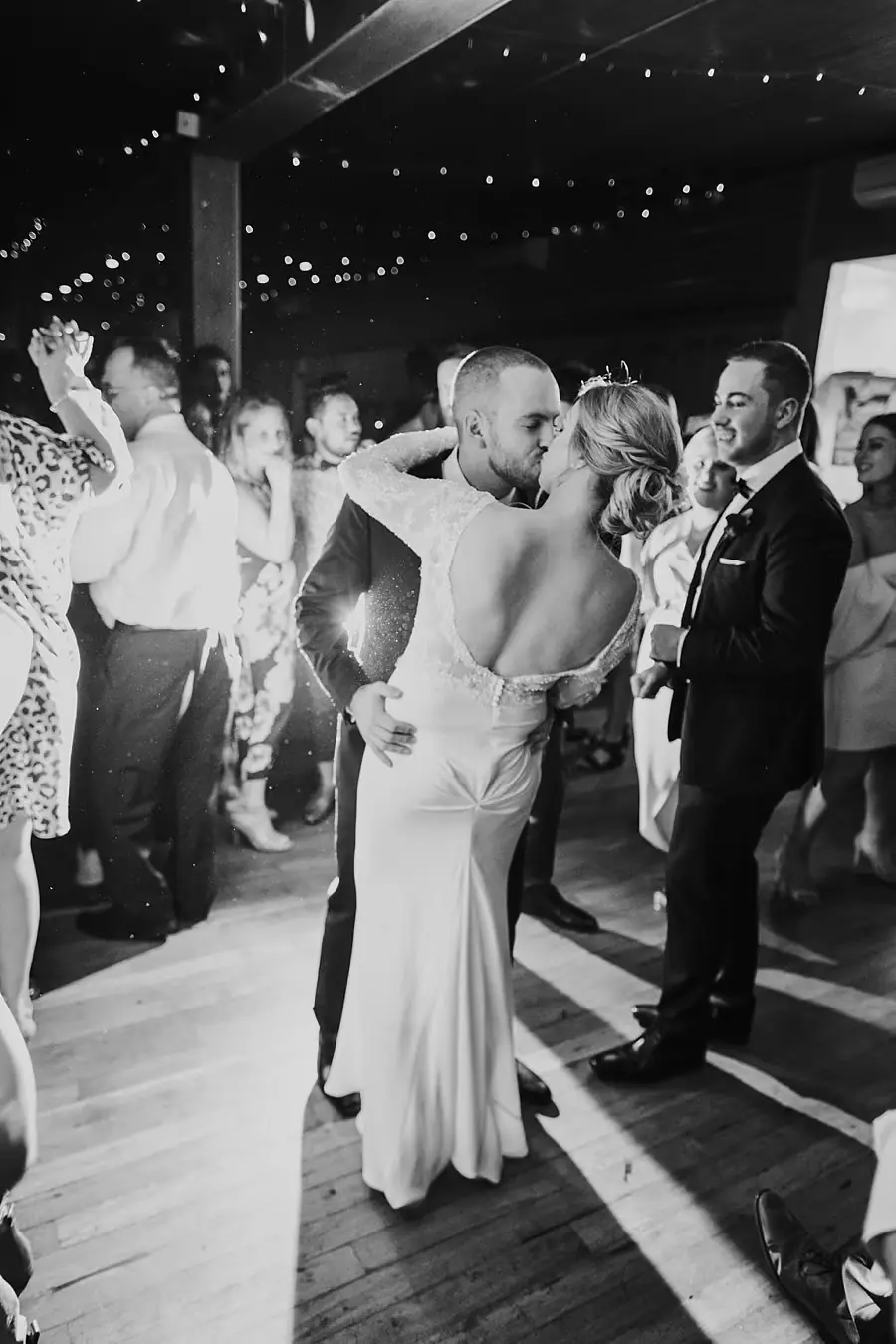 Black-and-white photograph of the bride dancing with guests beneath string lights.