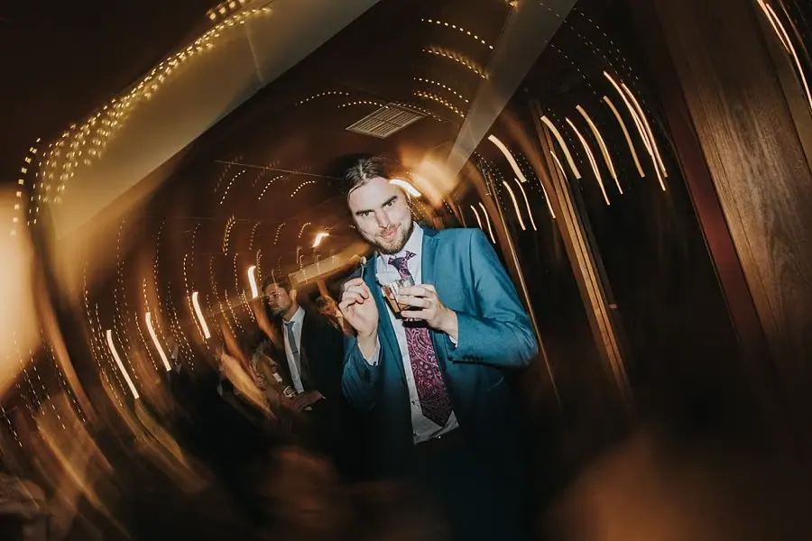 Man in a blue suit holding a drink and posing beneath blurred reception lights.