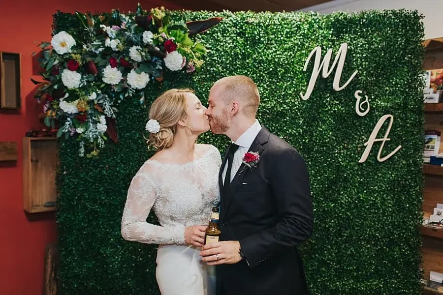 Bride and groom kissing in front of a green floral photo backdrop.