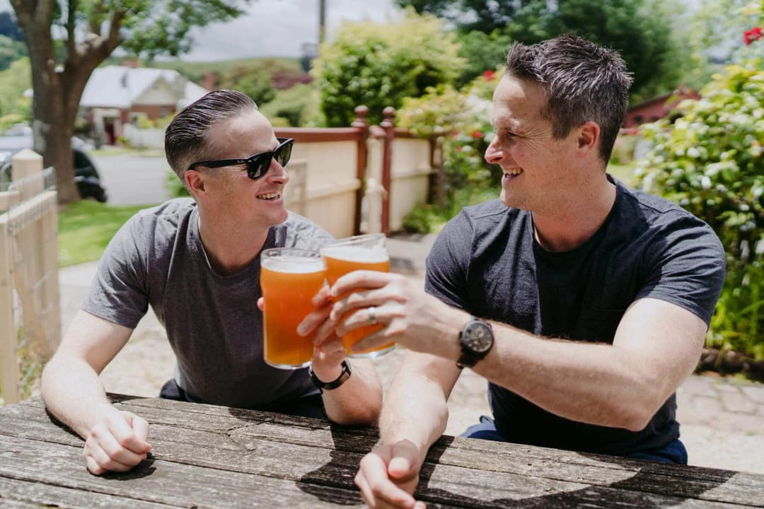 Two men clinking beer glasses at an outdoor wooden table.