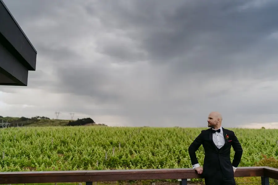 Groom leaning beside a timber railing overlooking vineyard rows beneath storm clouds.