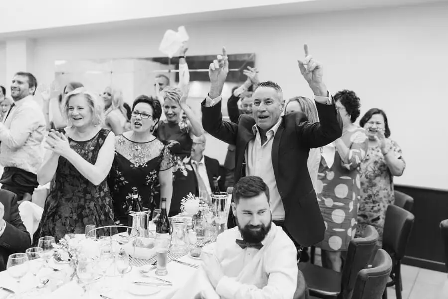 Black-and-white photograph of guests cheering with raised glasses around a reception table.