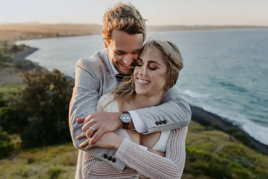 Groom embracing the smiling bride from behind beside a coastal cliff.
