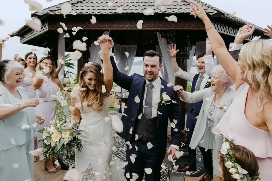 Couple raising their joined hands beneath a shower of white petals.