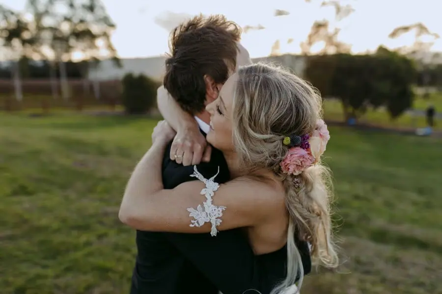 Bride hugging a man in a dark suit in a sunlit field.