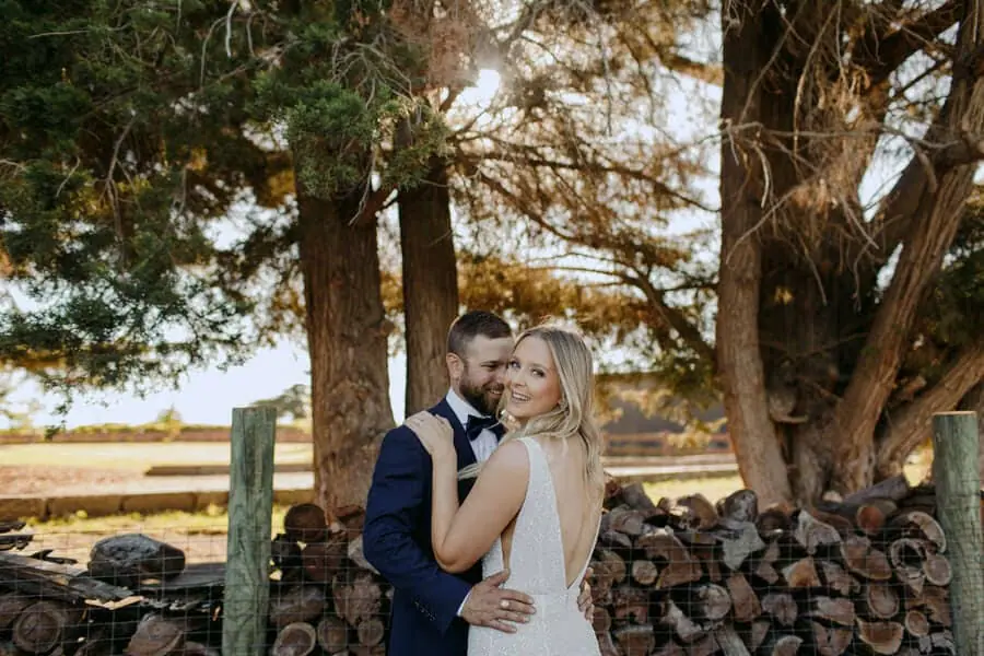 Bride and groom embracing beside stacked logs beneath tall trees.