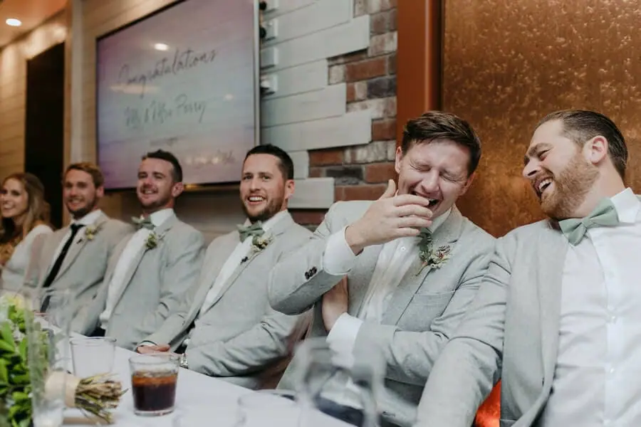 Men laughing together in pale suits at a reception table.