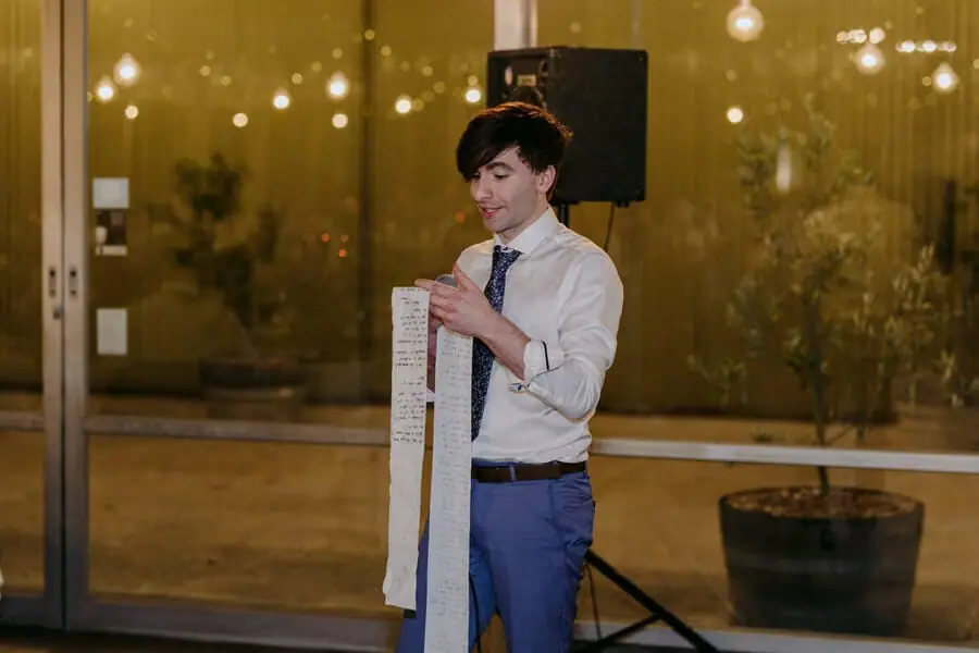 Man speaking while holding a long strip of speech notes beneath warm lights.