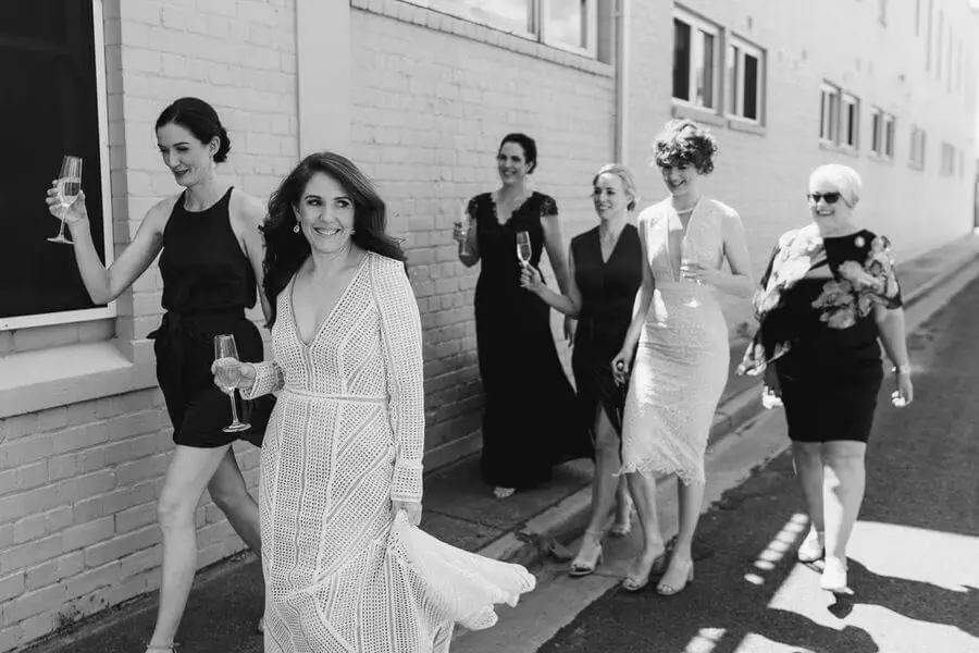 Black-and-white photograph of women walking beside a brick building with drinks.