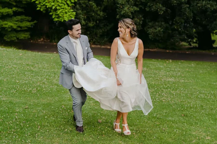 Groom lifting the bride's dress skirt as they laugh on a lawn.
