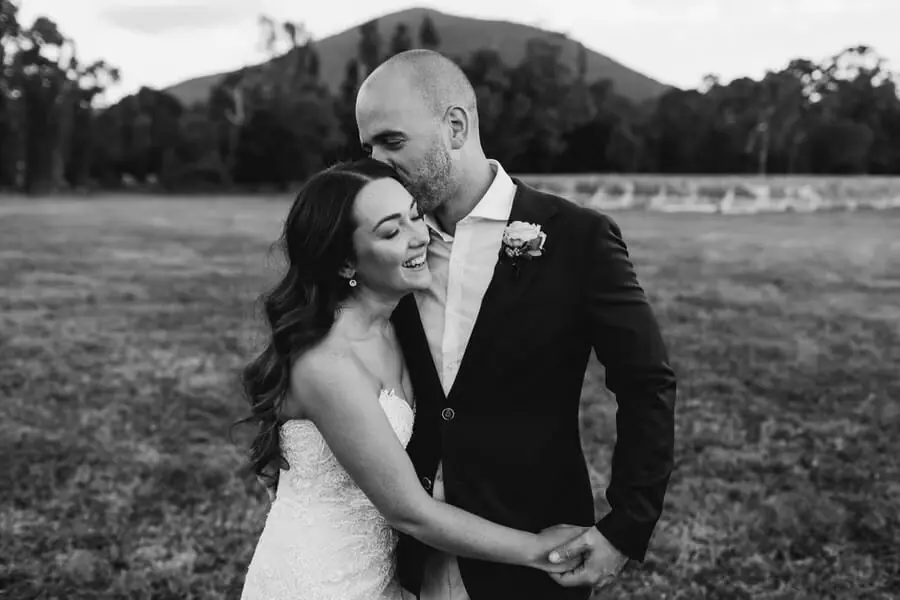 Black-and-white photograph of a groom kissing the bride's forehead in a field.