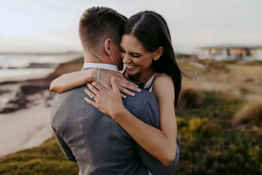 Bride smiling while embracing the groom beside coastal cliffs.