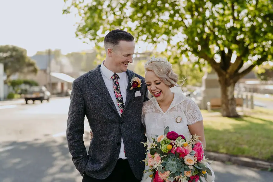 Bride and groom smiling together beneath a tree with her colourful bouquet.