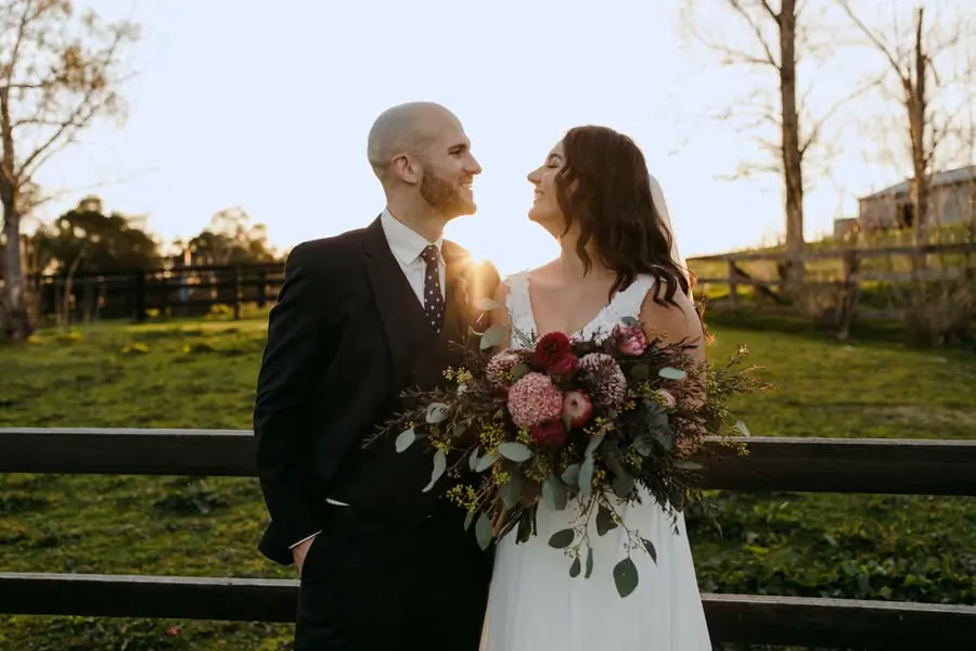 Couple smiling at each other beside a timber fence in evening light.