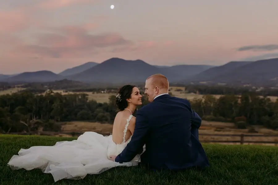 Couple sitting on a grassy hill beneath mountains and a pink sunset sky.