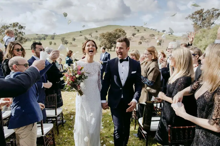 Bride and groom laughing as guests throw white petals over them.