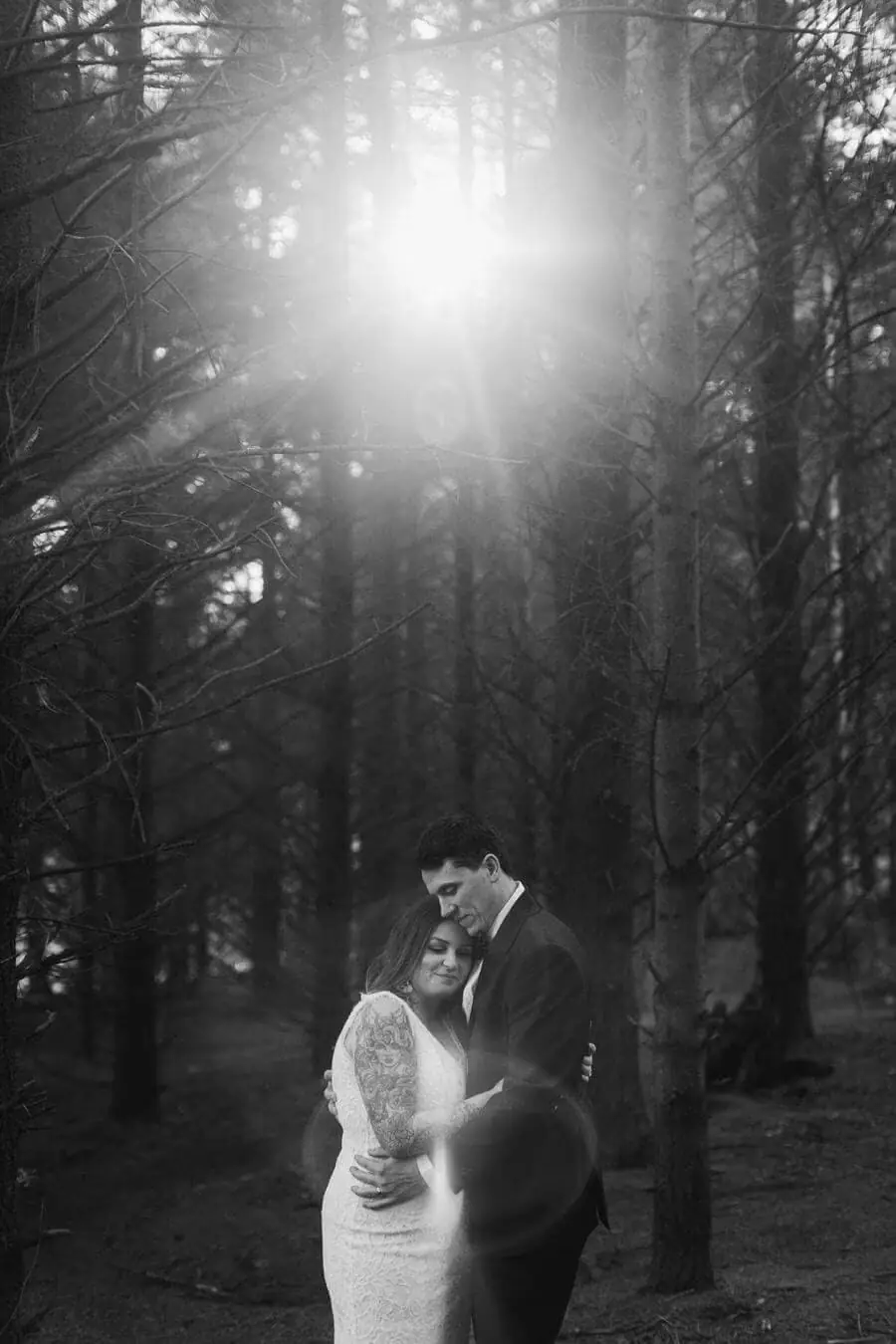 Black-and-white portrait of a couple embracing beneath sunlit forest trees.
