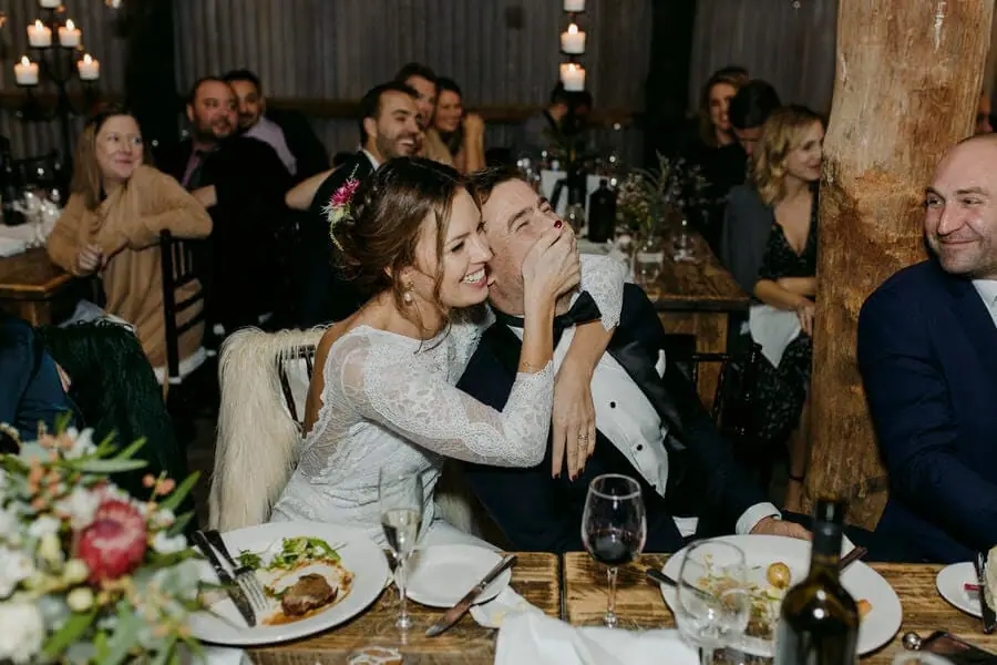 Bride kissing the groom's cheek while seated at a reception table.