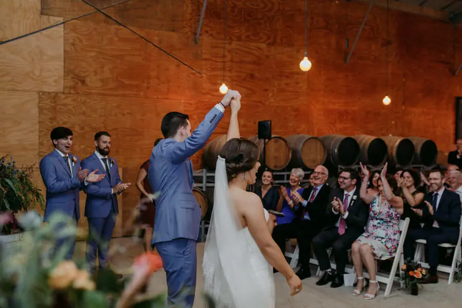 Groom raising the bride's hand as guests applaud in a barrel-lined ceremony room.